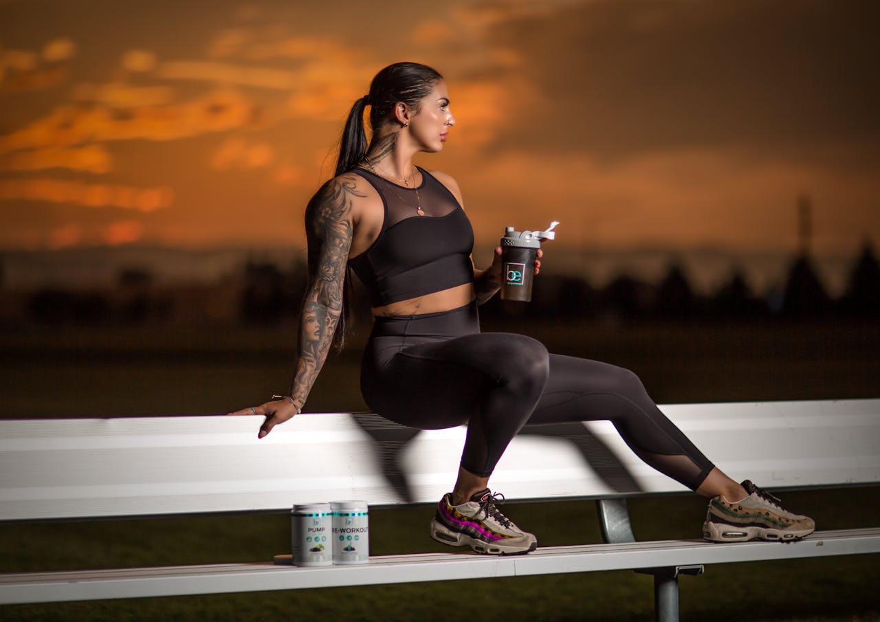 Athletic woman in activewear sits on a bench, enjoying sunset with a tumbler in hand.