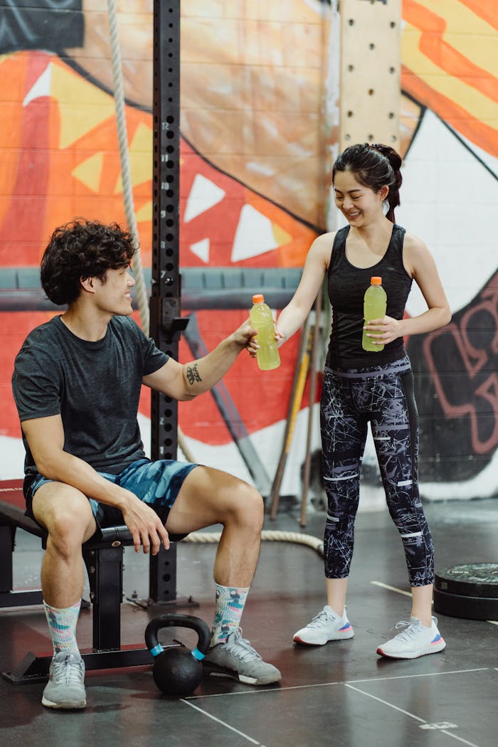 An Asian couple in activewear exchanging drinks in a gym setting, smiling and happy.